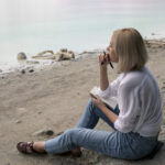 Beautiful blonde girl sits on the shore of the lake and writes in a notebook.
